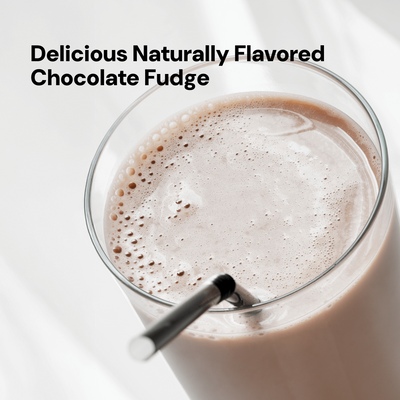 Overhead view of a clear glass containing a frothy, light brown drink labeled 'Delicious Naturally Flavored Chocolate Fudge'. A black and white striped straw is in the glass. The background is a bright, white surface.